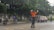 A traffic officer wearing an orange uniform stands in the middle of a road with arms raised, likely directing traffic. Nearby, a red car and several motorcyclists are visible. The background is lined with lush green trees and a fence.