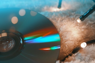A CD or DVD reflecting vibrant colors of light sits on a soft, fluffy surface. Small holiday string lights with a warm glow are intertwined nearby, casting a cozy and festive atmosphere.