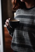 A person holding a dark coffee cup, wearing a patterned sweater. The focus is on the cup with frothy coffee, and the setting appears cozy, with soft lighting.