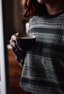 A person holding a dark coffee cup, wearing a patterned sweater. The focus is on the cup with frothy coffee, and the setting appears cozy, with soft lighting.