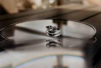 Two silver rings are elegantly placed on a reflective surface, capturing a soft and intimate atmosphere. The background is blurred, accentuating the focus on the rings and the subtle reflections they create.
