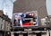 A large billboard on the side of a building displays images of several people in a collage, with text that reads 'A Europe of Equals'. The surrounding environment includes a typical European street with buildings and a pub below the billboard. The outdoor setting features cloudy skies and some bare tree branches.