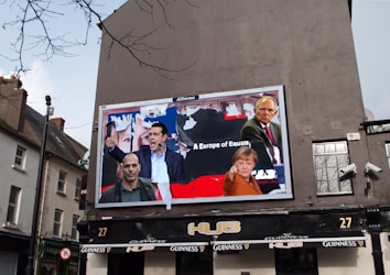 A large billboard on the side of a building displays images of several people in a collage, with text that reads 'A Europe of Equals'. The surrounding environment includes a typical European street with buildings and a pub below the billboard. The outdoor setting features cloudy skies and some bare tree branches.