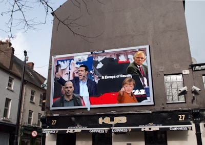 A large billboard on the side of a building displays images of several people in a collage, with text that reads 'A Europe of Equals'. The surrounding environment includes a typical European street with buildings and a pub below the billboard. The outdoor setting features cloudy skies and some bare tree branches.