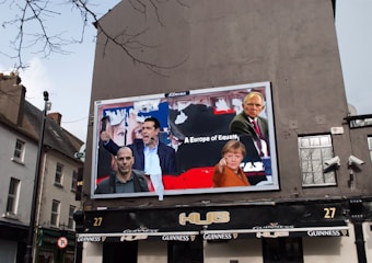 A large billboard on the side of a building displays images of several people in a collage, with text that reads 'A Europe of Equals'. The surrounding environment includes a typical European street with buildings and a pub below the billboard. The outdoor setting features cloudy skies and some bare tree branches.