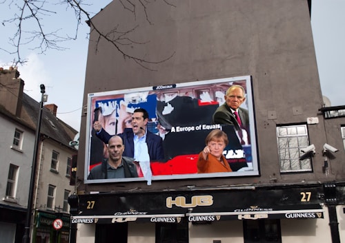 A large billboard on the side of a building displays images of several people in a collage, with text that reads 'A Europe of Equals'. The surrounding environment includes a typical European street with buildings and a pub below the billboard. The outdoor setting features cloudy skies and some bare tree branches.
