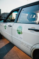A close-up view of a white car with a sticker on the side door featuring the number 24 and some text with an automotive theme. The car has a retro design, and the sunlight creates a warm glow on the surface.
