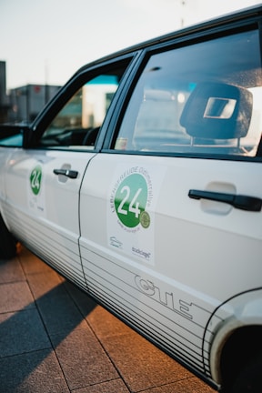 A close-up view of a white car with a sticker on the side door featuring the number 24 and some text with an automotive theme. The car has a retro design, and the sunlight creates a warm glow on the surface.