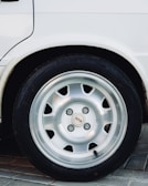 A close-up view of a car wheel featuring a silver alloy rim and a black tire. The image includes part of a white vehicle's body and captures the wheel resting on a paved surface.