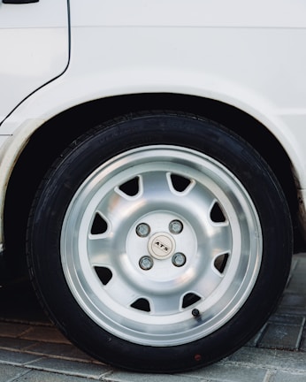 A close-up view of a car wheel featuring a silver alloy rim and a black tire. The image includes part of a white vehicle's body and captures the wheel resting on a paved surface.