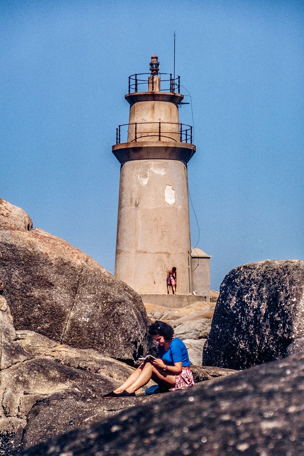 woman in blue shirt sitting on rock near white lighthouse under blue sky during daytime