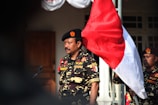 A man in a military-style uniform stands outdoors, holding a large red and white flag. The uniform features various patches and a beret with a distinctive emblem. Another person in similar attire is partially visible in the background.