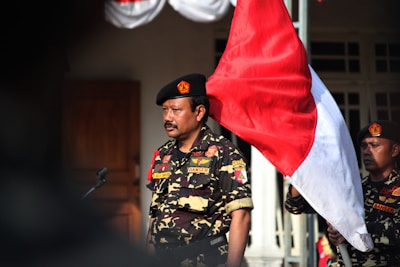 A man in a military-style uniform stands outdoors, holding a large red and white flag. The uniform features various patches and a beret with a distinctive emblem. Another person in similar attire is partially visible in the background.