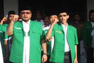 Several men are standing together, wearing green jackets and black hats. They appear to be in an outdoor setting, possibly participating in a gathering or event. Their expressions and raised fists suggest a sense of unity or protest.