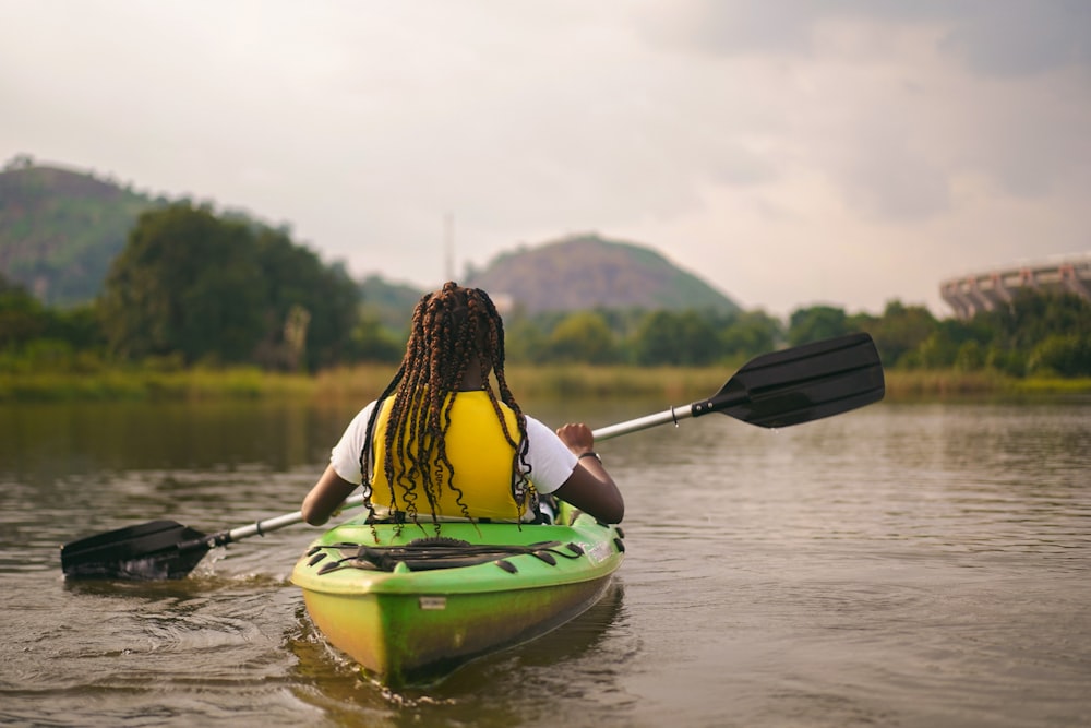 woman in yellow and black stripe tank top sitting on green kayak on lake during daytime