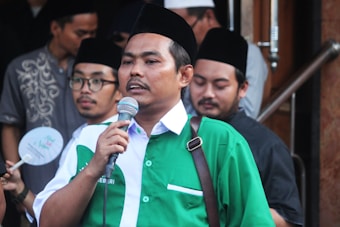 A person wearing a green and white shirt speaks into a microphone while standing outdoors. Several other individuals are present in the background, some wearing black traditional caps.