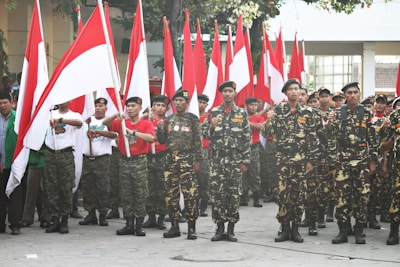 A group of uniformed individuals stand in formation, holding red and white flags, which are often associated with national symbols. The uniforms have a military-style appearance with camouflage patterns. The setting appears to be outdoors with a building and trees in the background.