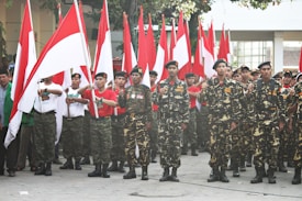 A group of uniformed individuals stand in formation, holding red and white flags, which are often associated with national symbols. The uniforms have a military-style appearance with camouflage patterns. The setting appears to be outdoors with a building and trees in the background.