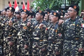 A group of individuals in uniform stand together in formation, raising their fists in a gesture of solidarity or chanting. The uniforms are camouflage-patterned with black berets, and they have patches with various insignia. Red and white flags are visible in the background.