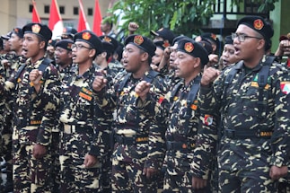 A group of individuals in uniform stand together in formation, raising their fists in a gesture of solidarity or chanting. The uniforms are camouflage-patterned with black berets, and they have patches with various insignia. Red and white flags are visible in the background.