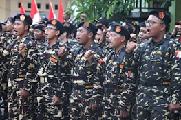 A group of individuals in uniform stand together in formation, raising their fists in a gesture of solidarity or chanting. The uniforms are camouflage-patterned with black berets, and they have patches with various insignia. Red and white flags are visible in the background.