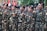 A group of individuals in uniform stand together in formation, raising their fists in a gesture of solidarity or chanting. The uniforms are camouflage-patterned with black berets, and they have patches with various insignia. Red and white flags are visible in the background.