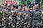 A group of individuals in uniform stand together in formation, raising their fists in a gesture of solidarity or chanting. The uniforms are camouflage-patterned with black berets, and they have patches with various insignia. Red and white flags are visible in the background.