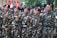 A group of individuals in uniform stand together in formation, raising their fists in a gesture of solidarity or chanting. The uniforms are camouflage-patterned with black berets, and they have patches with various insignia. Red and white flags are visible in the background.