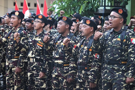 A group of individuals in uniform stand together in formation, raising their fists in a gesture of solidarity or chanting. The uniforms are camouflage-patterned with black berets, and they have patches with various insignia. Red and white flags are visible in the background.