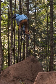 A person is mountain biking through a forest, airborne after jumping off a dirt ramp. The surroundings are dense woods with tall trees, creating a natural and adventurous setting.