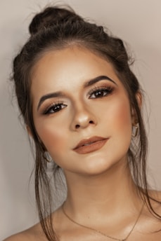 A close-up portrait of a young woman with styled hair in a bun. Her makeup is expertly applied, emphasizing her eyes with bold eyelashes and perfectly shaped eyebrows. Her skin has a smooth texture, with a natural glow. The background is a soft, neutral tone.