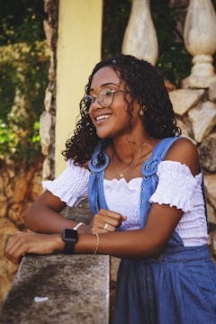 A woman with curly hair and glasses is smiling while leaning on a stone railing. She is wearing a white off-the-shoulder top and a blue denim dress with decorative flower details. An outdoor setting with rustic stone walls and lush greenery forms the background.
