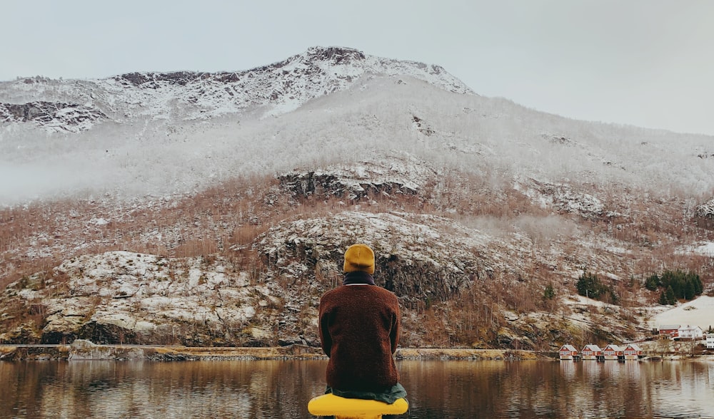 person in red hoodie and yellow knit cap sitting on rock facing snow covered mountain during