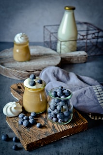A rustic arrangement of a small glass jar filled with fresh blueberries and another jar with a creamy dessert topped with blueberries. A meringue sits beside them on a wooden board, with a cloth napkin nearby. In the background, there is a bottle of milk inside a metal wire basket, placed on a wooden tray.