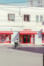 A person is riding a bicycle in front of a commercial building with red and white branding. The building features several posters and the name Ooredoo above a glass door. The street is empty, and the day is clear.