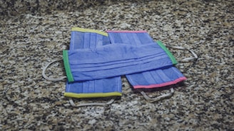 Four colorful surgical masks are arranged on a speckled granite surface. Each mask features a different colored trim: green, yellow, and pink, complementing their blue fabric.