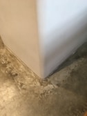 A close-up view of a corner where a smooth, white wall meets a textured concrete floor. The floor has a mottled appearance with various shades of gray and beige, giving it a somewhat industrial look.