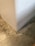 A close-up view of a corner where a smooth, white wall meets a textured concrete floor. The floor has a mottled appearance with various shades of gray and beige, giving it a somewhat industrial look.