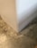 A close-up view of a corner where a smooth, white wall meets a textured concrete floor. The floor has a mottled appearance with various shades of gray and beige, giving it a somewhat industrial look.