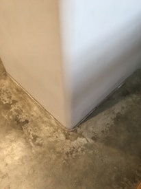A close-up view of a corner where a smooth, white wall meets a textured concrete floor. The floor has a mottled appearance with various shades of gray and beige, giving it a somewhat industrial look.