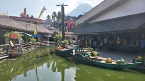 A scenic outdoor marketplace with wooden shops and tropical decor is situated along a green, calm waterway. Fruit-laden boats are docked beside a wooden pier. Brightly colored buildings and palm trees are in the background, with a mountain peak visible behind them.