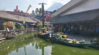 A scenic outdoor marketplace with wooden shops and tropical decor is situated along a green, calm waterway. Fruit-laden boats are docked beside a wooden pier. Brightly colored buildings and palm trees are in the background, with a mountain peak visible behind them.