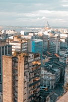 An urban landscape with a cluster of high-rise buildings, some showing signs of wear and aging. The foreground highlights a partially abandoned building with exposed floors and vegetation on the roof. In the background, a mixture of modern and older architectural styles is visible, along with industrial cranes near the waterfront.