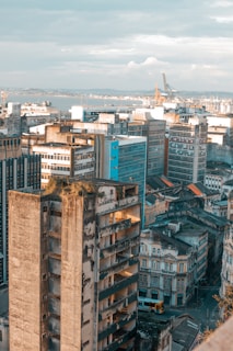 An urban landscape with a cluster of high-rise buildings, some showing signs of wear and aging. The foreground highlights a partially abandoned building with exposed floors and vegetation on the roof. In the background, a mixture of modern and older architectural styles is visible, along with industrial cranes near the waterfront.
