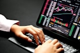 Hands typing on a laptop keyboard with a screen displaying financial graphs and stock market data. Multiple charts and numbers are visible, indicating online trading or financial analysis.