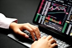 Hands typing on a laptop keyboard with a screen displaying financial graphs and stock market data. Multiple charts and numbers are visible, indicating online trading or financial analysis.