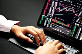 Hands typing on a laptop keyboard with a screen displaying financial graphs and stock market data. Multiple charts and numbers are visible, indicating online trading or financial analysis.