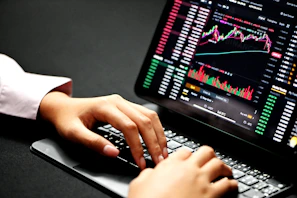 Close-up of hands typing on a laptop with financial graphs on the screen.