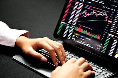 Hands typing on a laptop keyboard with a screen displaying financial graphs and stock market data. Multiple charts and numbers are visible, indicating online trading or financial analysis.