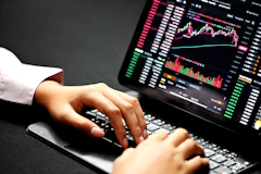 Close-up of hands typing on a laptop with financial graphs and trading data on the screen.